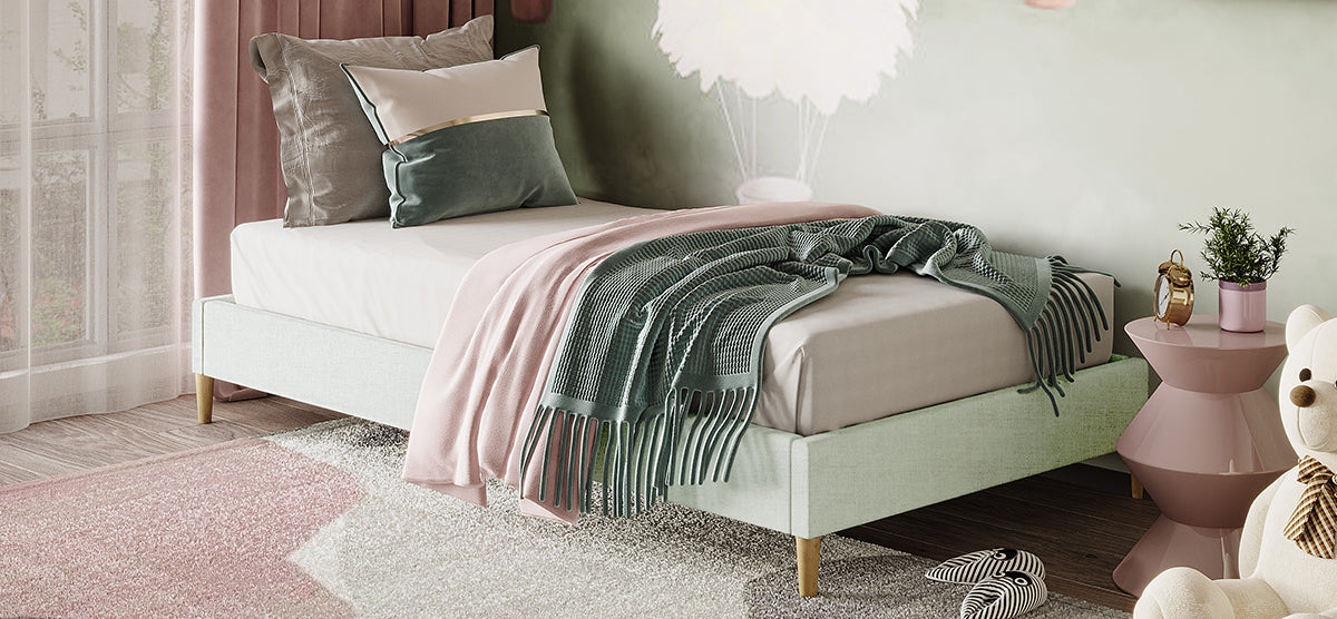 A pastel-themed bedroom corner featuring a modern platform bed with a pink throw and green knit blanket, accompanied by a pink side table with a plant and alarm clock.