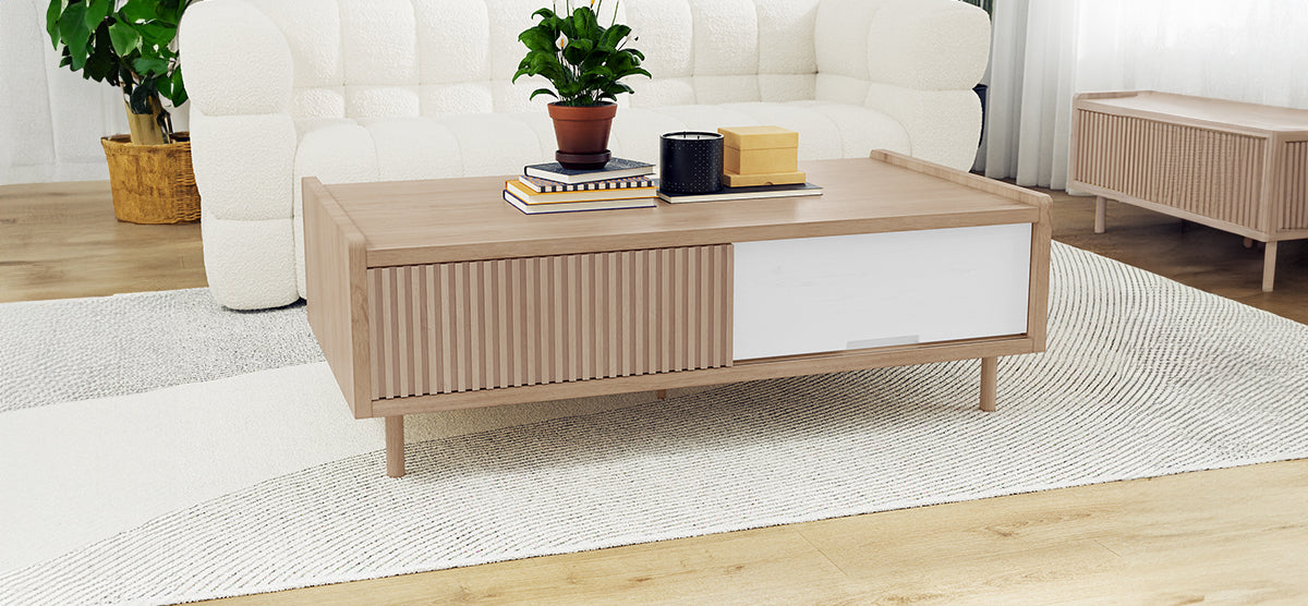 Modern living room with a low wooden coffee table featuring a potted plant, hardcover books, a black container, decorative boxes; white sofa and floor plant in background.