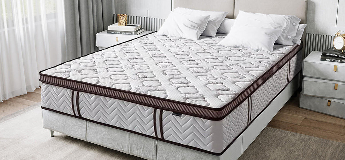A neatly made bed features a plush quilt-top mattress in white and light gray with dark trim, flanked by nightstands with books and decor.