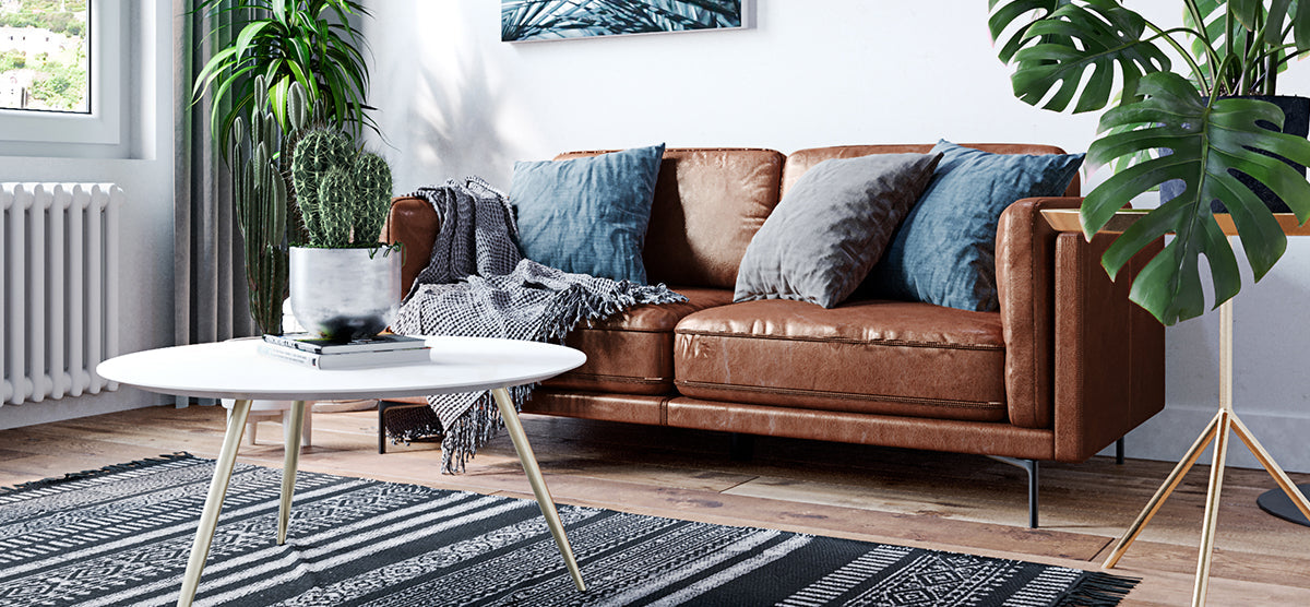 Modern living room corner featuring a round white coffee table with books and a cactus, a brown leather sofa, a black-and-white rug, and tall houseplants.