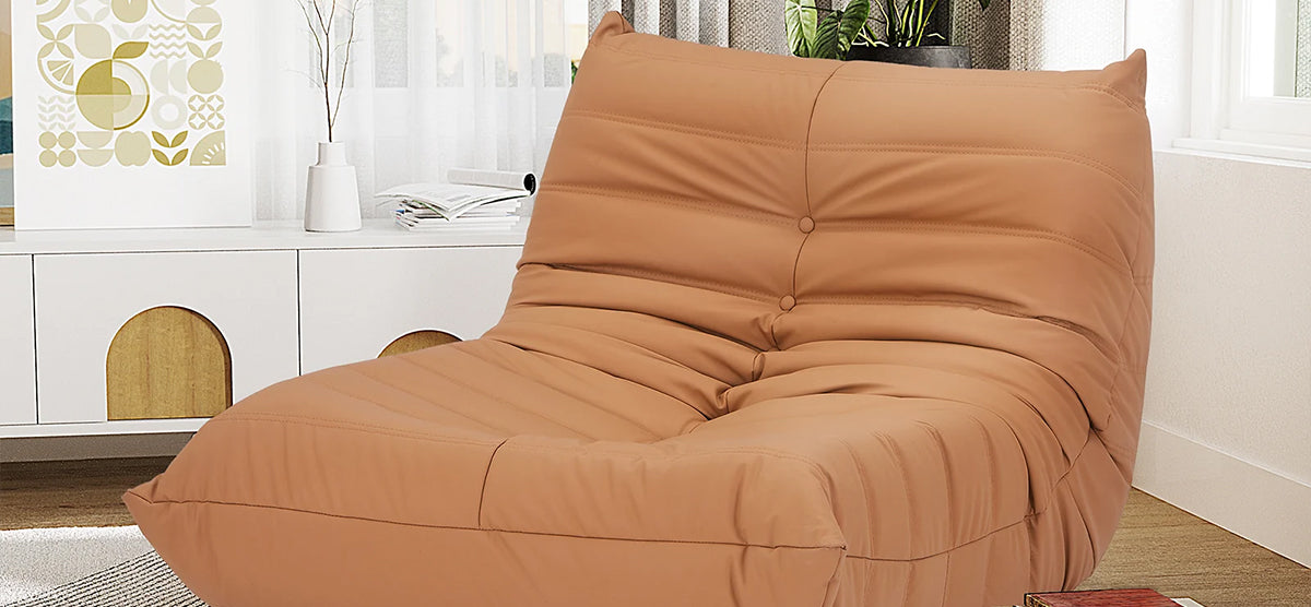 A warm tan faux-leather bean-bag chair on a light wood floor, near a white console with decorative items, framed geometric print, and a potted plant.