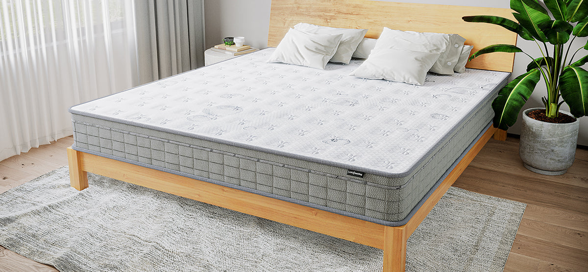 Minimalist bedroom with a light-wood platform bed, grey-bordered mattress, white quilted cover, two pillows, a bedside table with books and mug, sheer curtains, and potted plant.