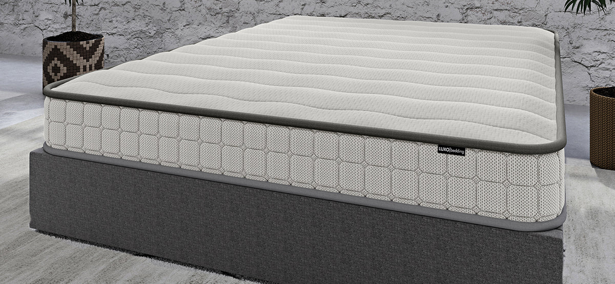 LUXObedding light-gray quilted mattress on dark gray upholstered base, featuring a grid-patterned side panel and softly stitched top, in a staged bedroom setting.