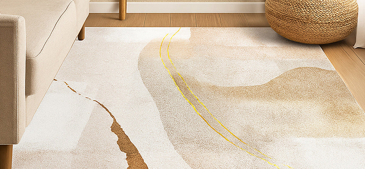 Abstract-patterned area rug in cream, tan, and sandy brown with gold accents on light-wood floor, near beige sofa and woven-fiber pouf.