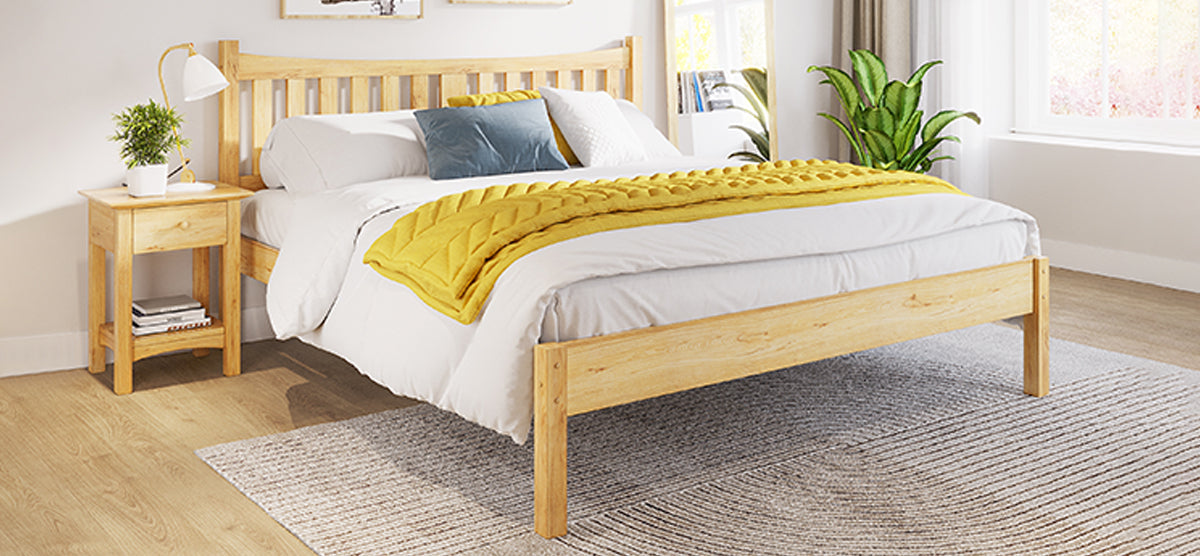 Minimal bedroom corner with a light wooden bed frame, white sheets, mustard throw, accent pillows, wooden nightstand, potted plant, table lamp, and a tall houseplant by a window.