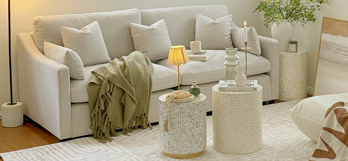 Neutral-toned living area with a cream upholstered sofa featuring throw pillows and an olive-green blanket. Two mosaic-tiled tables hold decorative objects, including a lamp, vase, and lit candle.