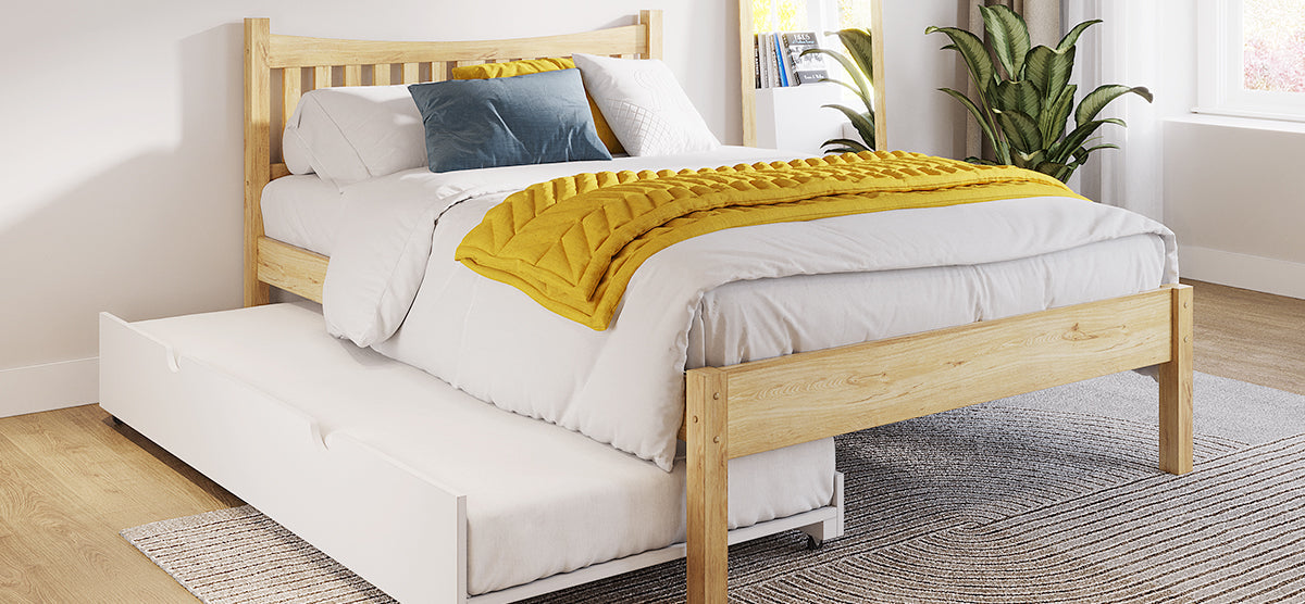 Wooden-frame bed with white linens, blue accent pillow, mustard quilt, and pull-out trundle. Light wood floor with rug, potted plant, and shelf with books in a bright, minimal bedroom.