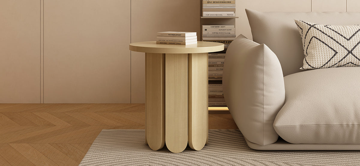 Minimalist living room with a light wood round side table featuring scalloped legs, stacked books, beige sofa with cushions, textured rug, and herringbone wood floor.