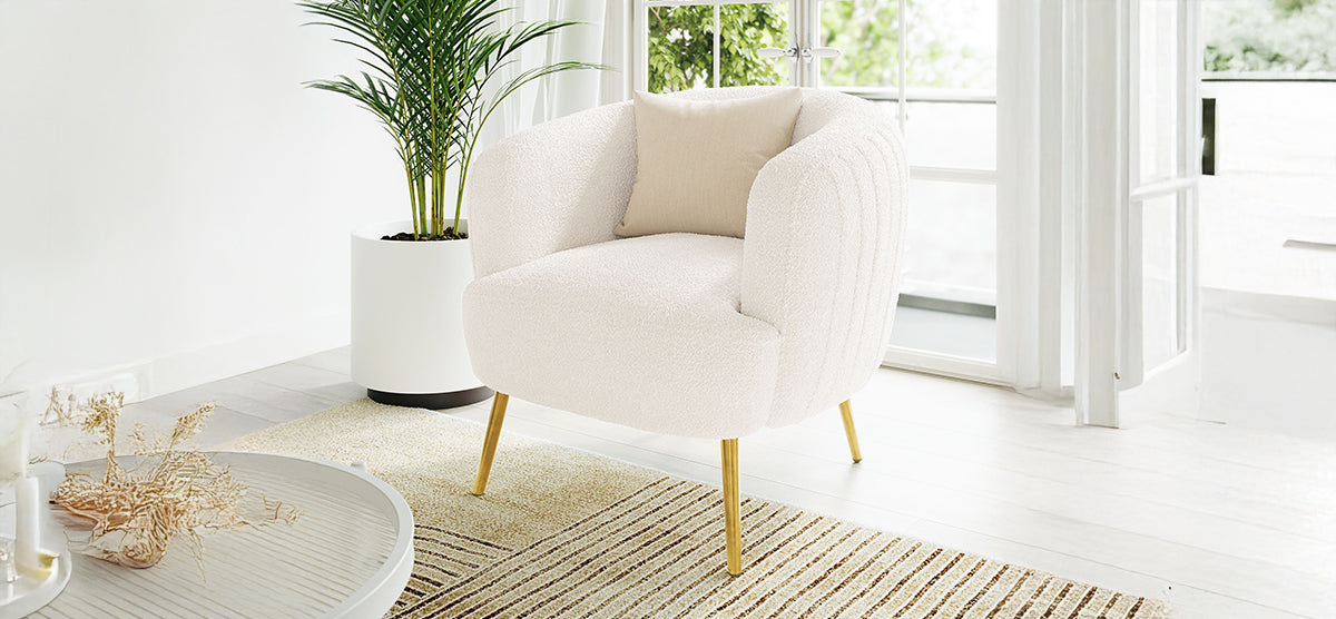 White boucle-upholstered armchair with gold legs and beige throw pillow beside a low coffee table with dried botanicals, set in a bright, airy room with glass doors.
