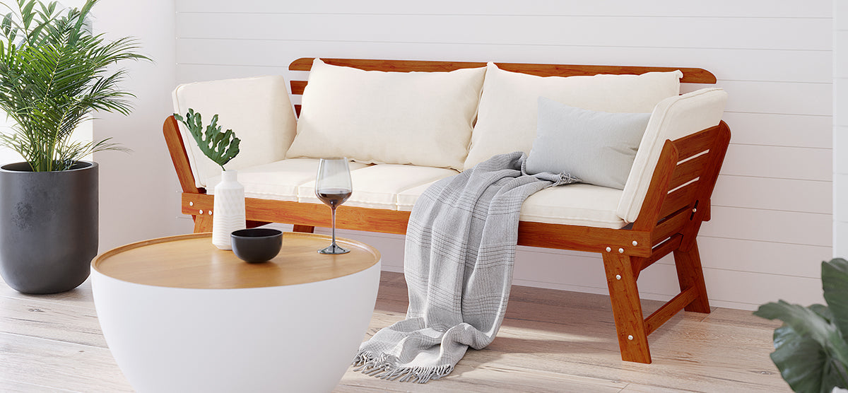 Minimalist seating area with a wooden-frame bench, off-white cushions, gray throw, round coffee table, white vase, single leaf, black bowl, wine glass, and potted plant.