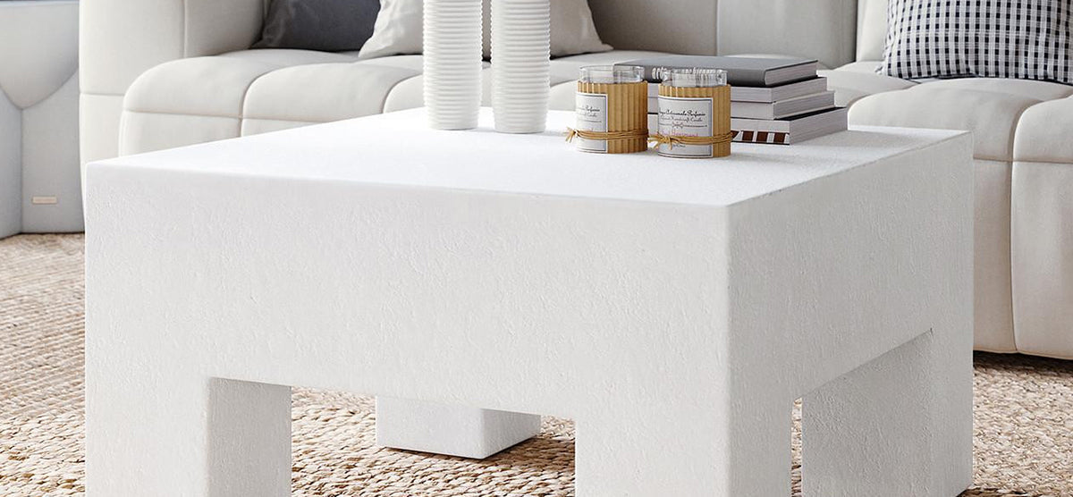 Modern living room with a chunky white coffee table featuring ribbed vases, candles, and books. Light sectional sofa and jute rug complete the minimalist vignette.