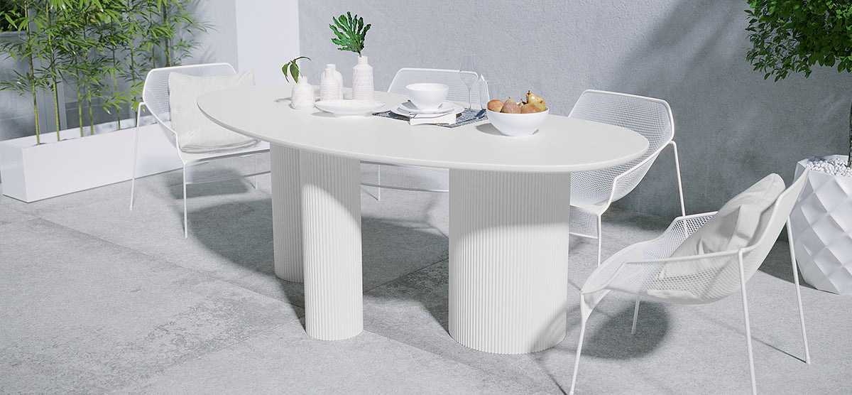 Minimalist outdoor dining setup featuring a white oval table with fluted legs, four wire-mesh chairs, decorative vases, and a bowl of pears, framed by bamboo and a potted tree.
