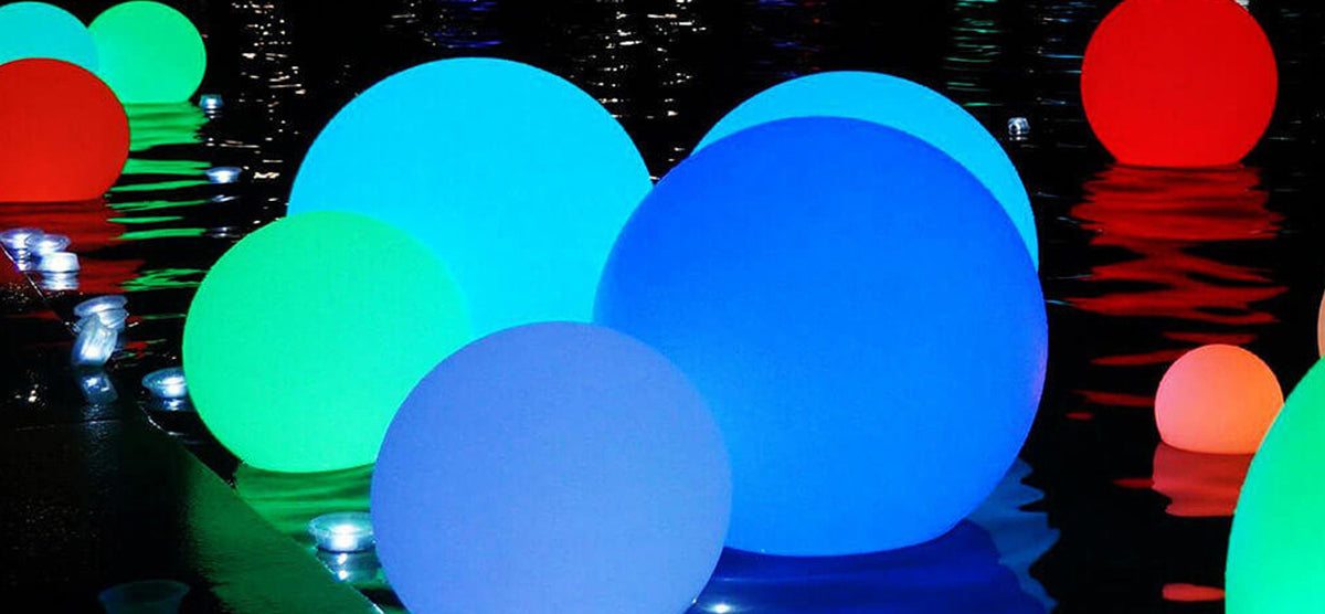 Glowing translucent spheres in blue, green, and red float on dark water, casting vibrant reflections, creating a festive, art-installation atmosphere.