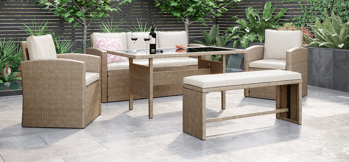 Modern patio with woven-rattan dining set featuring cushioned armchairs, a two-seat bench, and glass-top table with wine glasses; surrounded by lush greenery and a slat fence.