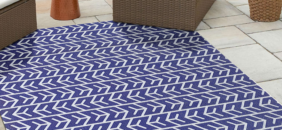 Outdoor seating area featuring a blue flat-weave rug with a white herringbone pattern, surrounded by brown wicker furniture, a copper side table, and a woven basket planter.