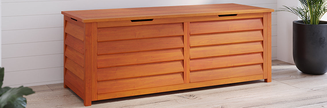 Rectangular wooden storage bench with slatted front and flat top, positioned indoors on a light-wood floor. A potted plant is partially visible on the right.