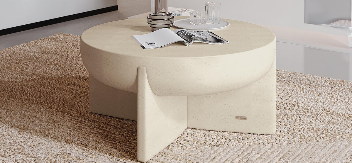 Low, round coffee table in cream with sculptural legs on a braided rug, featuring an open magazine, glassware on a tray, and a glass vase. Minimalist setting.