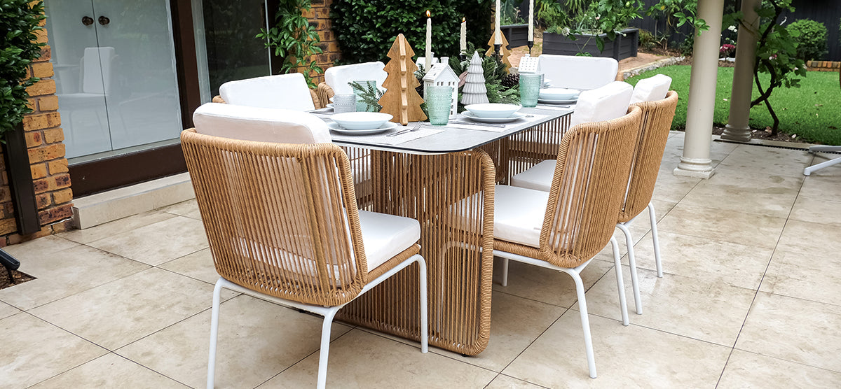 Outdoor dining setup on a tiled patio with a rectangular table, six rope-woven chairs, place settings, lit candles, and miniature decorative trees and houses.