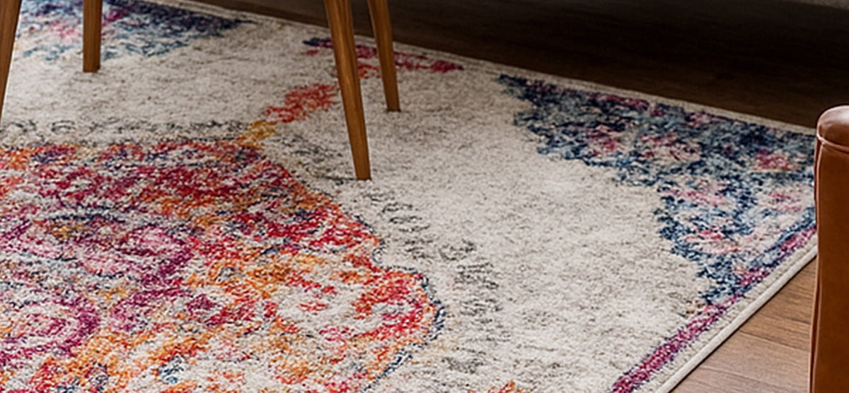 Close-up of a distressed multicolored area rug featuring pinks, oranges, and blues on a light background, with wooden furniture legs visible on a hardwood floor.