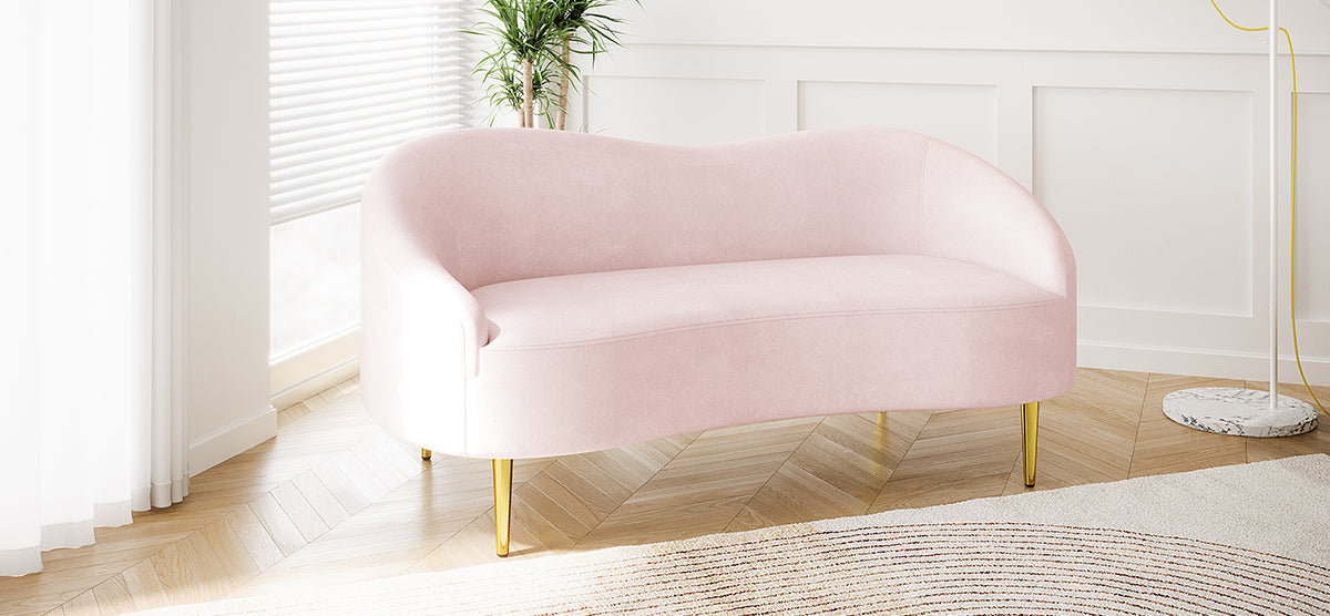 Blush-pink loveseat with gold-finished metal legs in a minimalist room, featuring white paneled walls, a tall leafy plant, and a slim floor lamp with a marble base.
