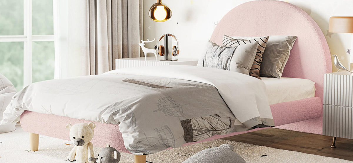 Softly styled bedroom corner with a low pink upholstered bed, light duvet, two plush toys, white nightstand with deer figurine, headphones, lamp, and sheer curtained window. Cozy ambiance.