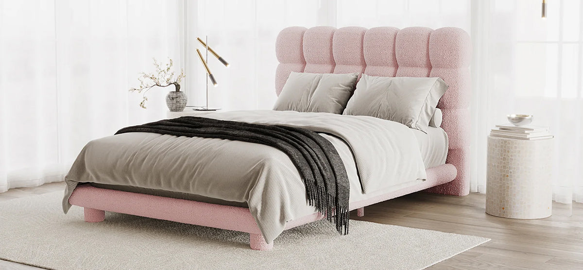 Modern bedroom corner with a low-profile pink upholstered bed, tufted headboard, light gray bedding, charcoal throw, sheer drapes, side table, decorative vase, and brass reading lamps.