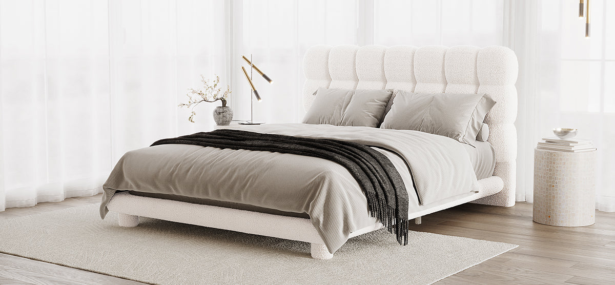 A serene bedroom corner featuring a low-profile bed with a white tufted headboard, neutral bedding, a charcoal throw, sheer curtains, a side table, and a brass reading lamp.