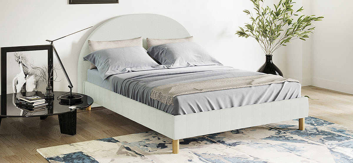 A modern bedroom corner featuring a low-profile platform bed with a rounded upholstered headboard, light gray bedding, a black side table with a lamp, books, and art, plus a tall black vase with greenery.