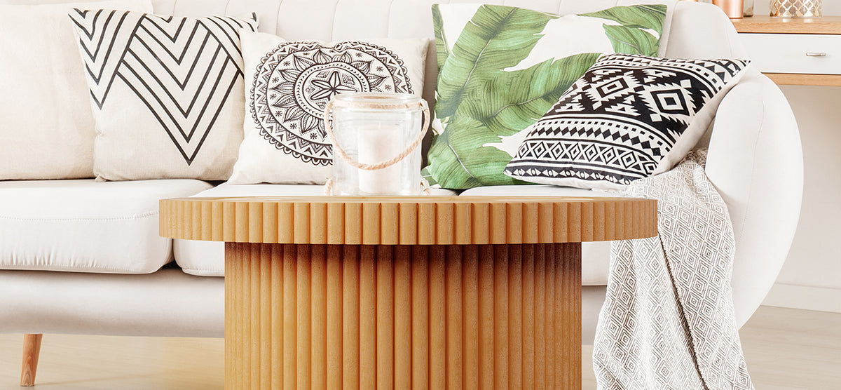 A cozy living-room vignette with a round wooden coffee table, upholstered sofa, geometric and botanical throw pillows, glass jar candle, and a patterned throw.