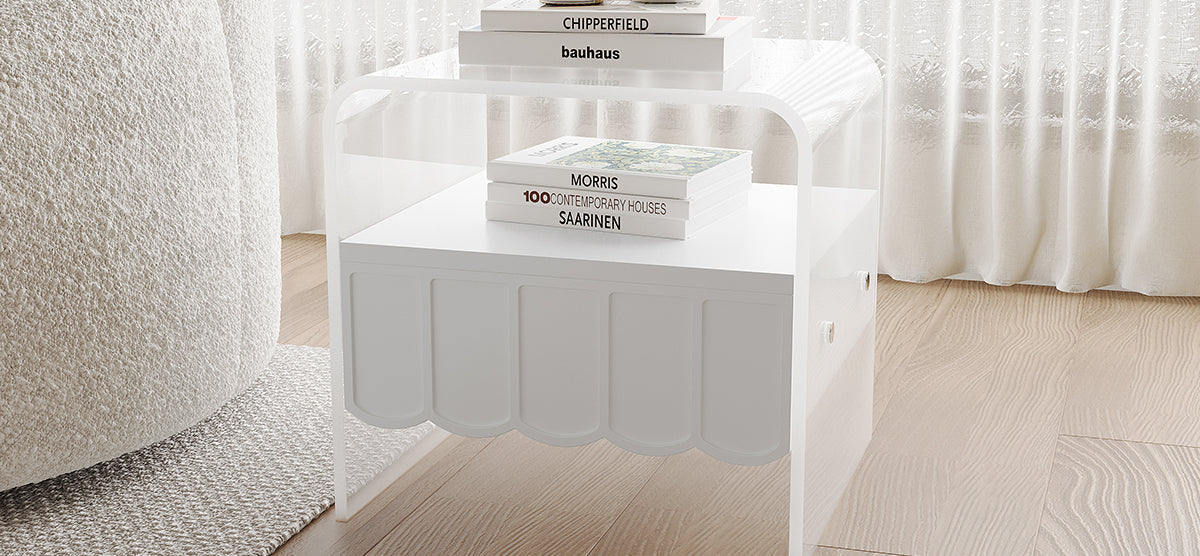 A minimalist living space corner with a white side table featuring clear acrylic sides, a matte white scalloped drawer, and art books. A cream boucle armchair partially sits on a textured rug.