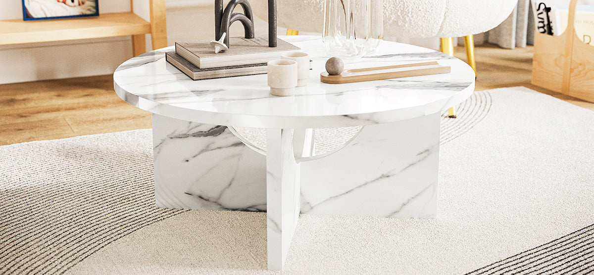 A small round white marble coffee table on a patterned rug, displaying books, a sculptural arch, cylindrical vases, a wooden board with a stone sphere, and a glass cloche.