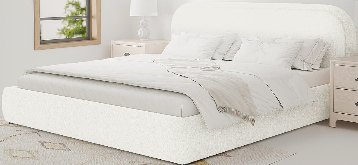 Minimal modern bedroom with a low-profile cream upholstered platform bed, white bedding, light-wood nightstands, a sculptural lamp, patterned rug, and window showing greenery.