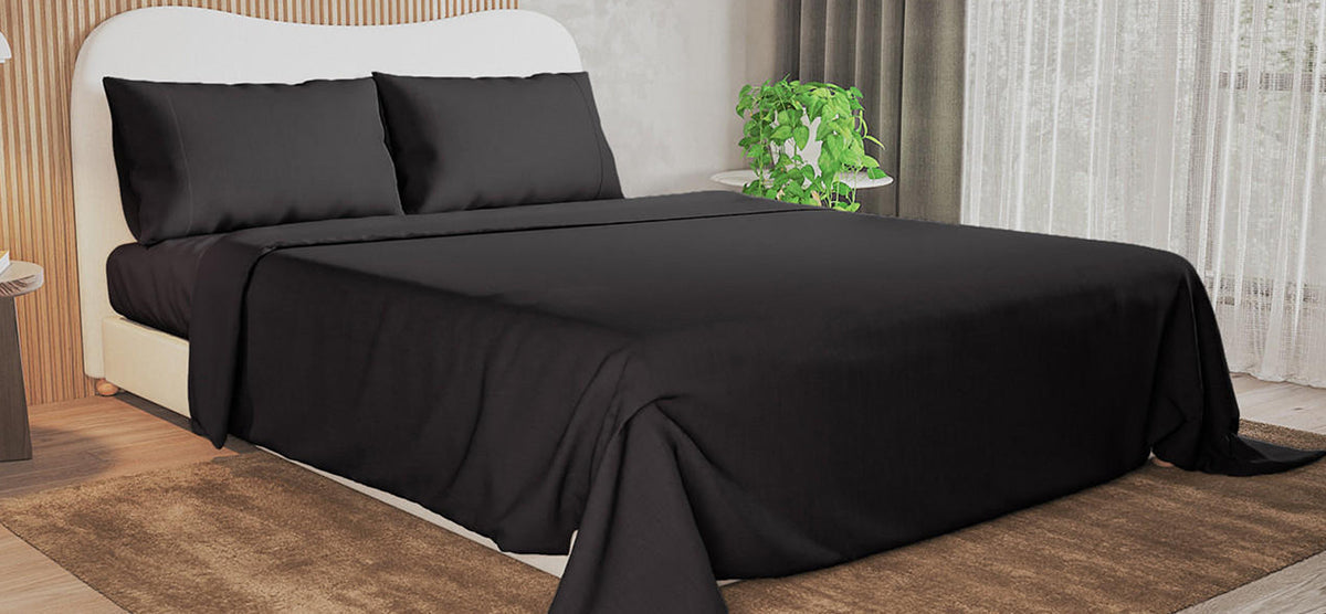 Modern bedroom with low-profile bed in black bedding, cream upholstered frame, wooden slat accent wall, side table with plant, sheer curtains, and medium-brown rug on wood floor.