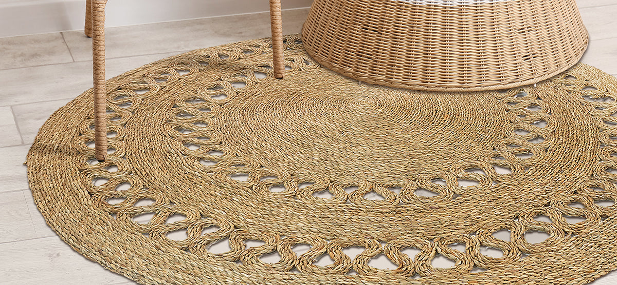 Round braided natural-fiber rug on floor with a woven wicker chair leg and wicker basket base resting on it, showcasing a rustic, textured decor style.