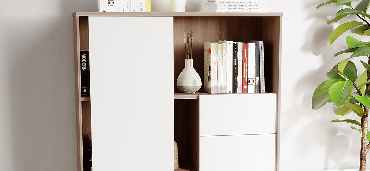 Modern light wood and white panel shelving unit with open shelves displaying a ceramic vase with dried twigs, books including “MODERN,” and two white drawers; accompanied by a tall leafy houseplant.
