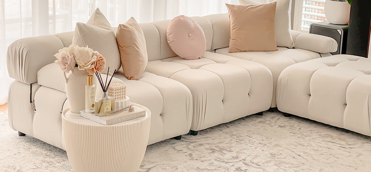 Cream-colored modular tufted sofa with blush and beige pillows, paired with a ribbed side table holding flowers, reed diffusers, a beaded candle, bottles, and a book.