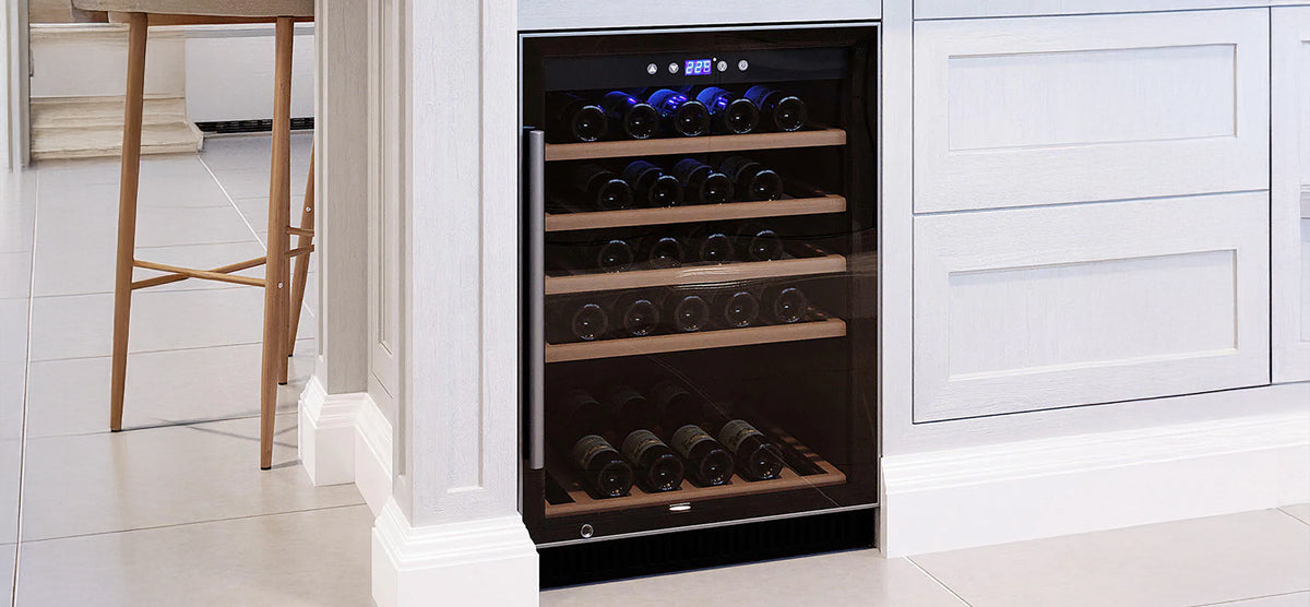 Built-in under-counter wine cooler integrated into white cabinetry, featuring a glass door, wooden shelves with wine bottles, and a digital temperature display; light-wood barstool visible nearby.