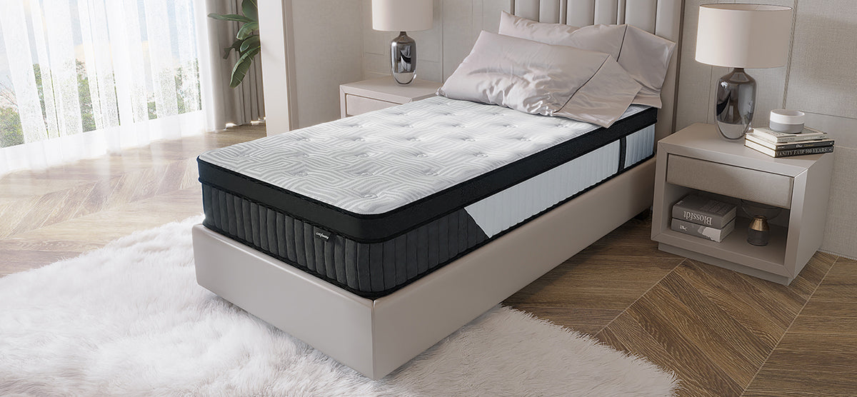 A modern bedroom corner featuring a twin-size upholstered platform bed with a pillow-top mattress, plush white rug, bedside table with lamp, books, and decorative item, alongside sheer curtains and a potted plant.