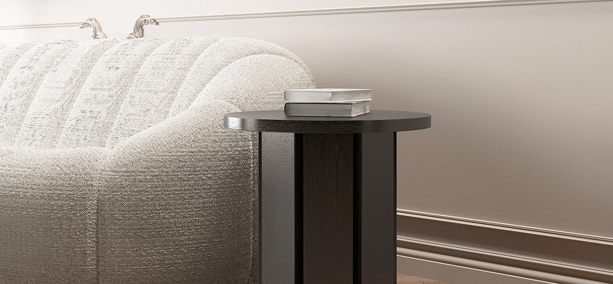 Cream-colored sofa arm next to a round black side table with two stacked hardcover books, set against a neutrally toned wall with simple molding.