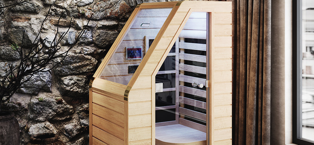 Indoor wooden infrared sauna pod with glass-front door, interior bench, and digital control panel. Features wood-slat construction with brass trim, set against a stone wall with a potted plant nearby.