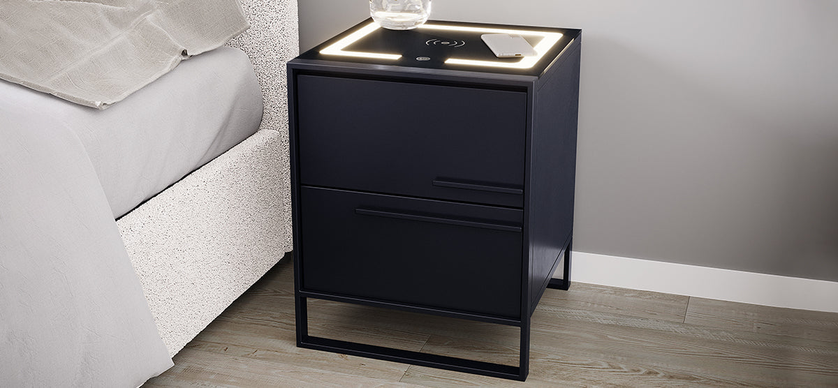 Modern bedside nightstand with dark finish, two drawers, raised metal base, and backlit top featuring wireless charging icon, smartphone, and glass carafe, next to upholstered bed.