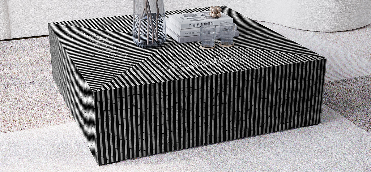 Square coffee table with a bold black-and-white striped pattern, featuring a vase with twigs, books, spherical sculpture, and ribbed glasses on coasters, on a textured rug.