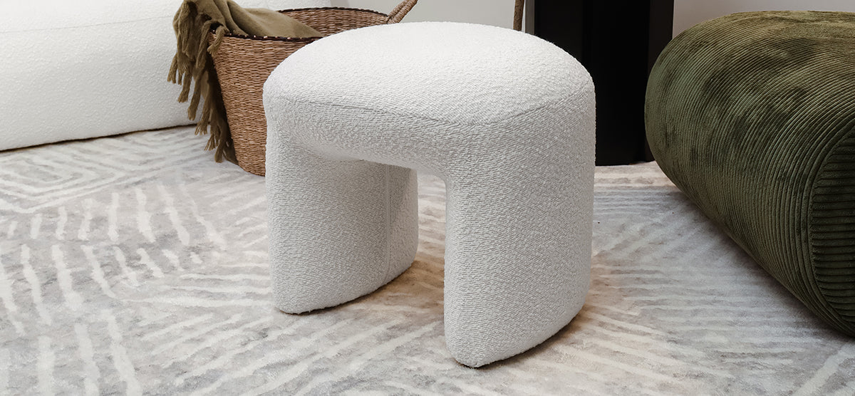 U-shaped modern upholstered stool in soft off-white boucle fabric, placed on a patterned area rug, with a woven basket and low olive-green ribbed bench in the background.