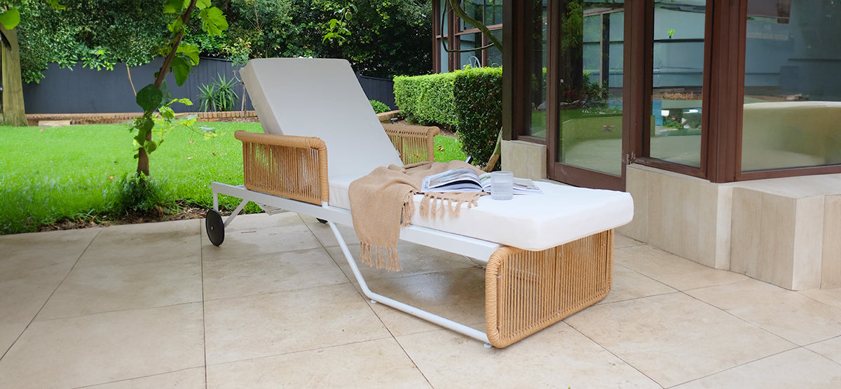 Outdoor terrace with a white-cushioned sun lounger featuring woven rattan panels, topped with a fringed throw, open book, and cup, set on neutral tiles.