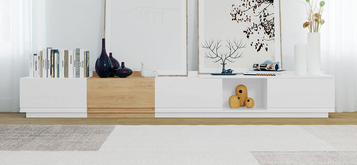 Modern sideboard with mixed wood and white panels, topped with books, dark vases, abstract sculpture, prints, antler-shaped rack, and tall vases with dried grasses. Minimalist decor.