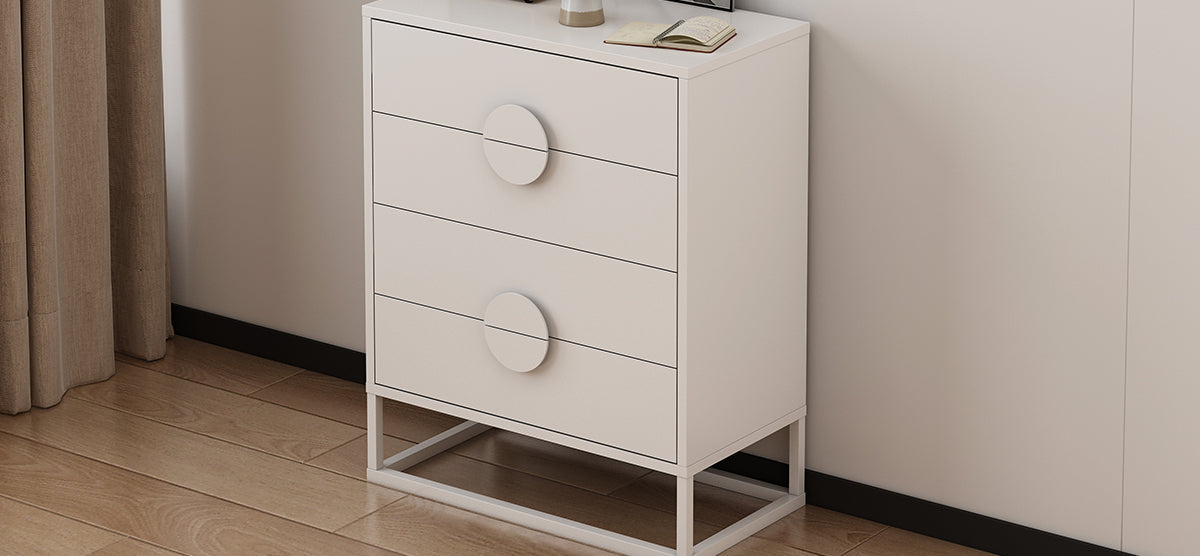 Modern white chest of drawers on slim metal frame with open notebook, small decorative vase or lamp, and framed picture on top, against a white wall.