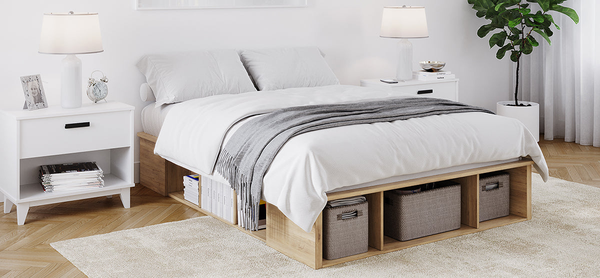 Modern bedroom with a low wooden platform bed featuring cubby storage. White linens, gray throw, white nightstands, lamps, books, framed photo, potted plant, and sheer-curtained window.