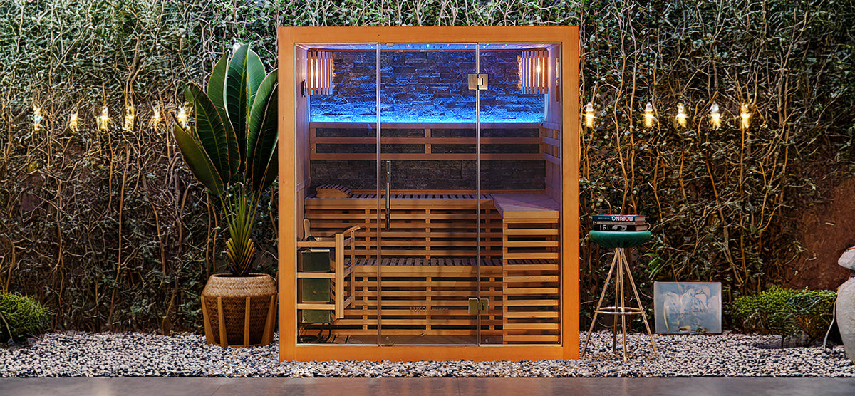 Outdoor sauna cube on white pebbles, featuring glass door, tiered wooden benches, blue LED lighting, large potted plant, string lights, stool with books, and a framed print.