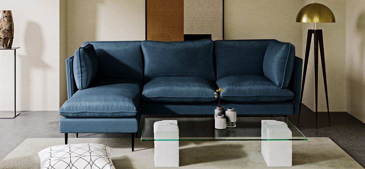 Modern living-room with deep-blue sectional sofa, glass-top coffee table, ceramic vases, area rug, white floor cushion, brass-toned floor lamp, and neutral wall art.