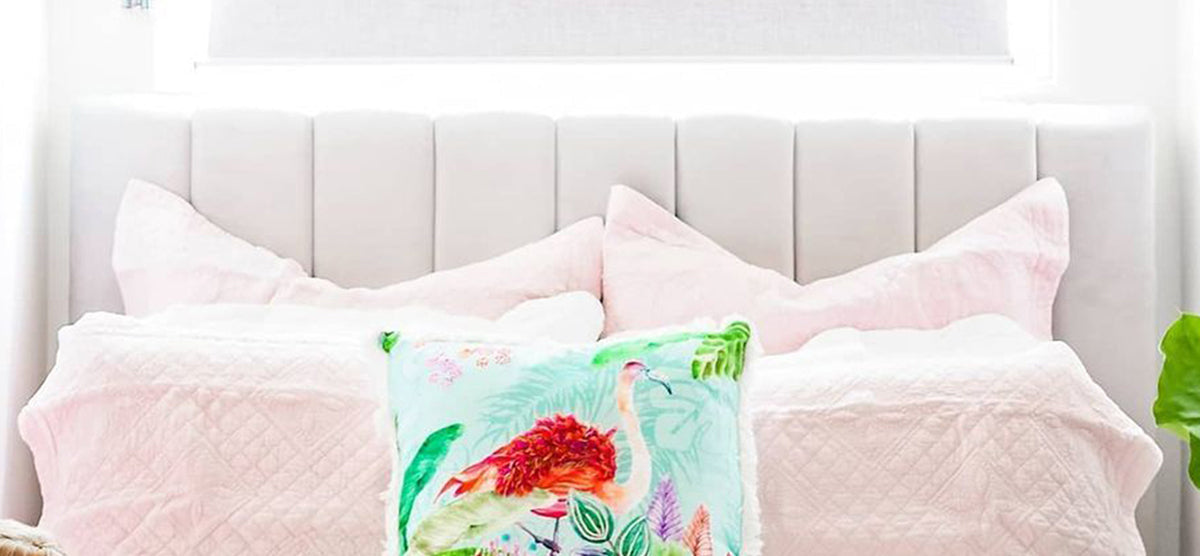 Neatly made bed with pastel pink bedding and a decorative throw cushion featuring a flamingo and tropical foliage. A green potted plant is partially visible beside the bed.